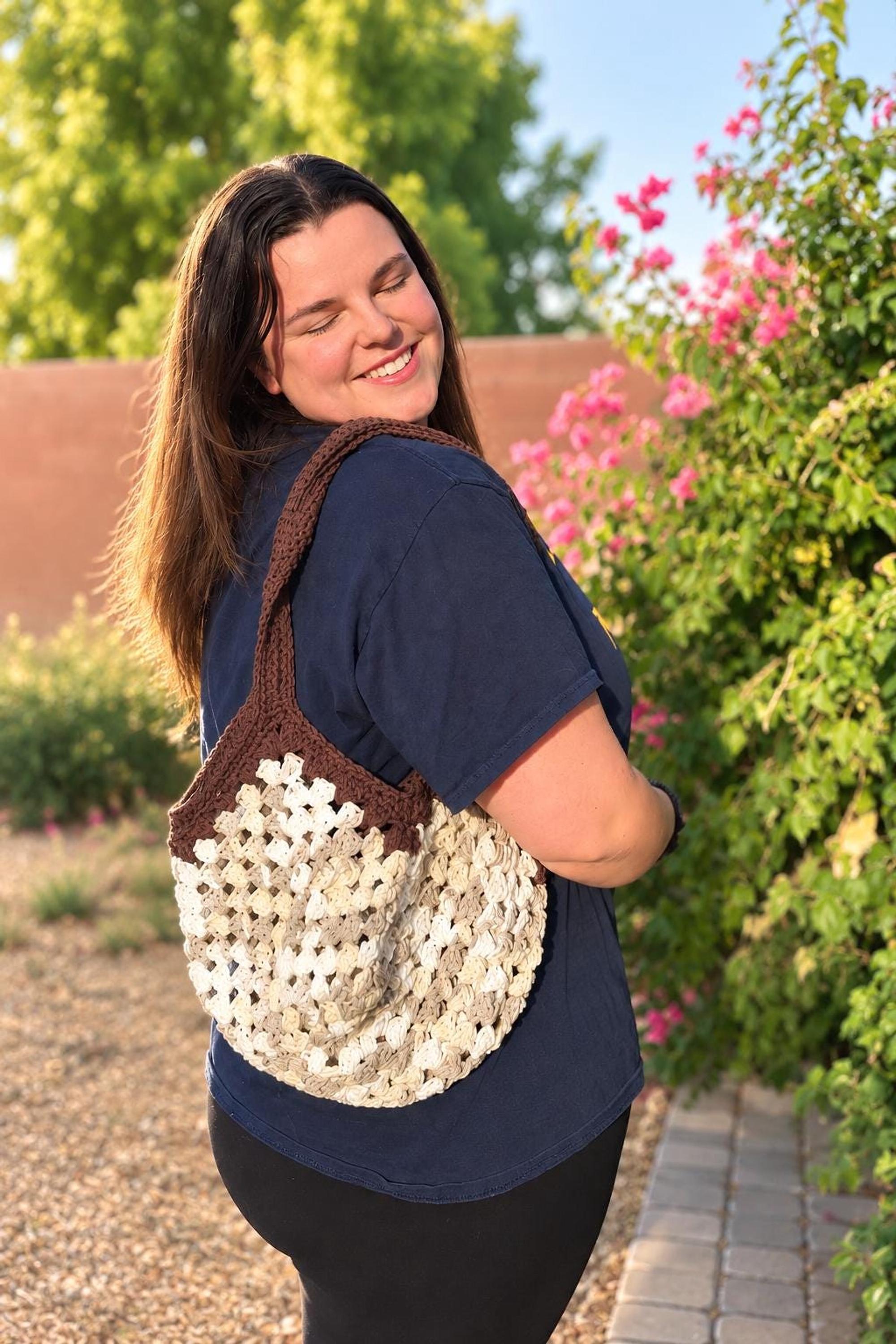 A smiling woman with long brown hair stands outdoors, wearing a navy t-shirt and black pants, with a round, off-white, brown, and tan crochet mesh bag hanging from her right shoulder.