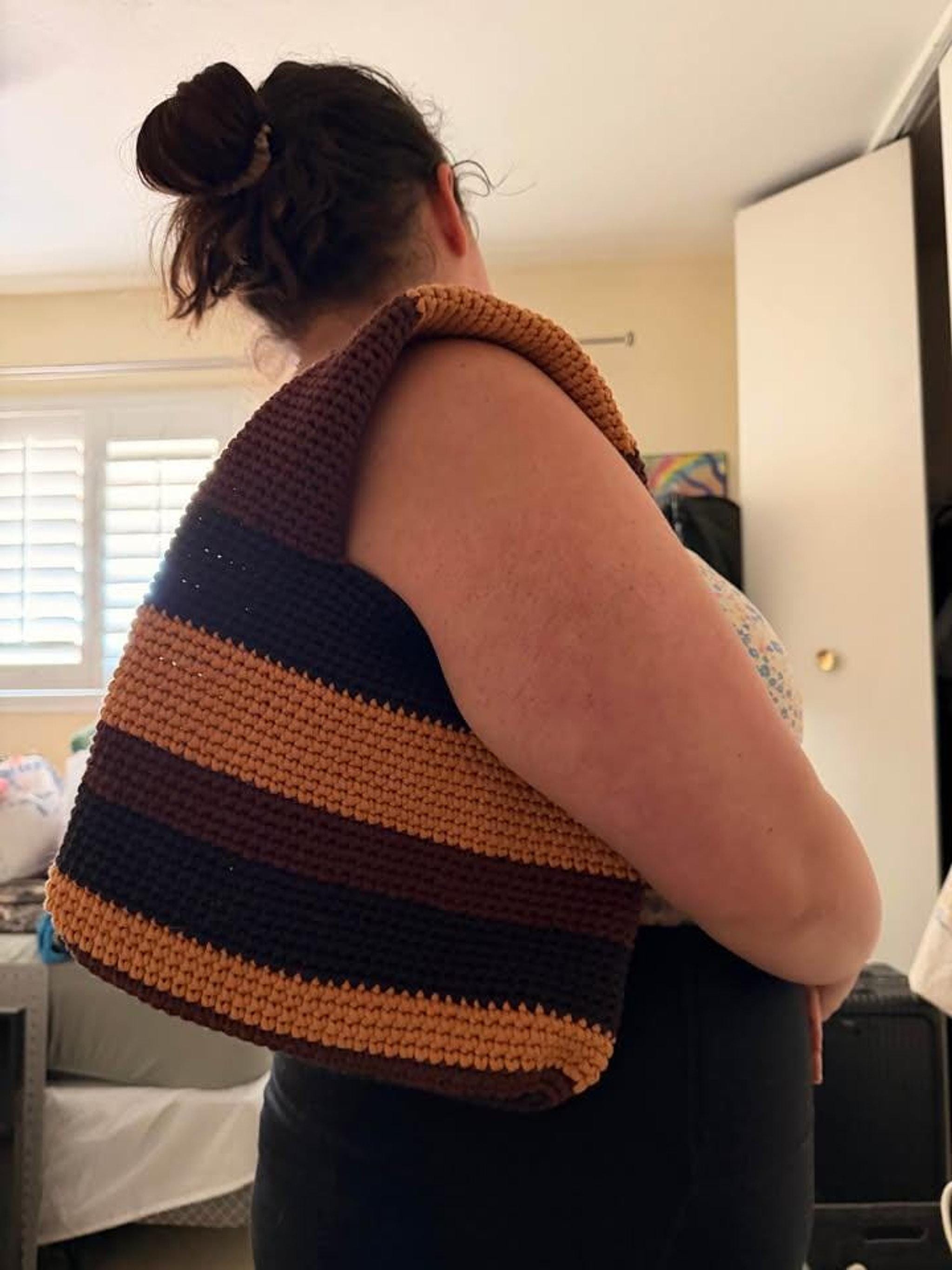 A person with brown hair in a bun wears a crocheted tote bag with horizontal stripes in dark brown, light brown, and orange over their right shoulder.