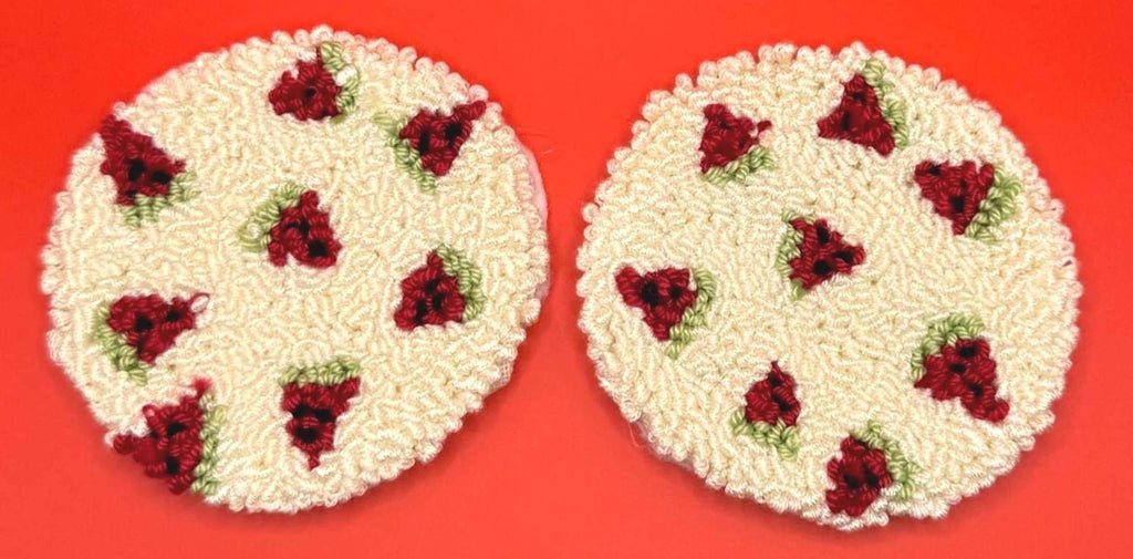 Two round punch needle coasters, each with a red strawberry pattern on a cream background, are shown on a red surface.