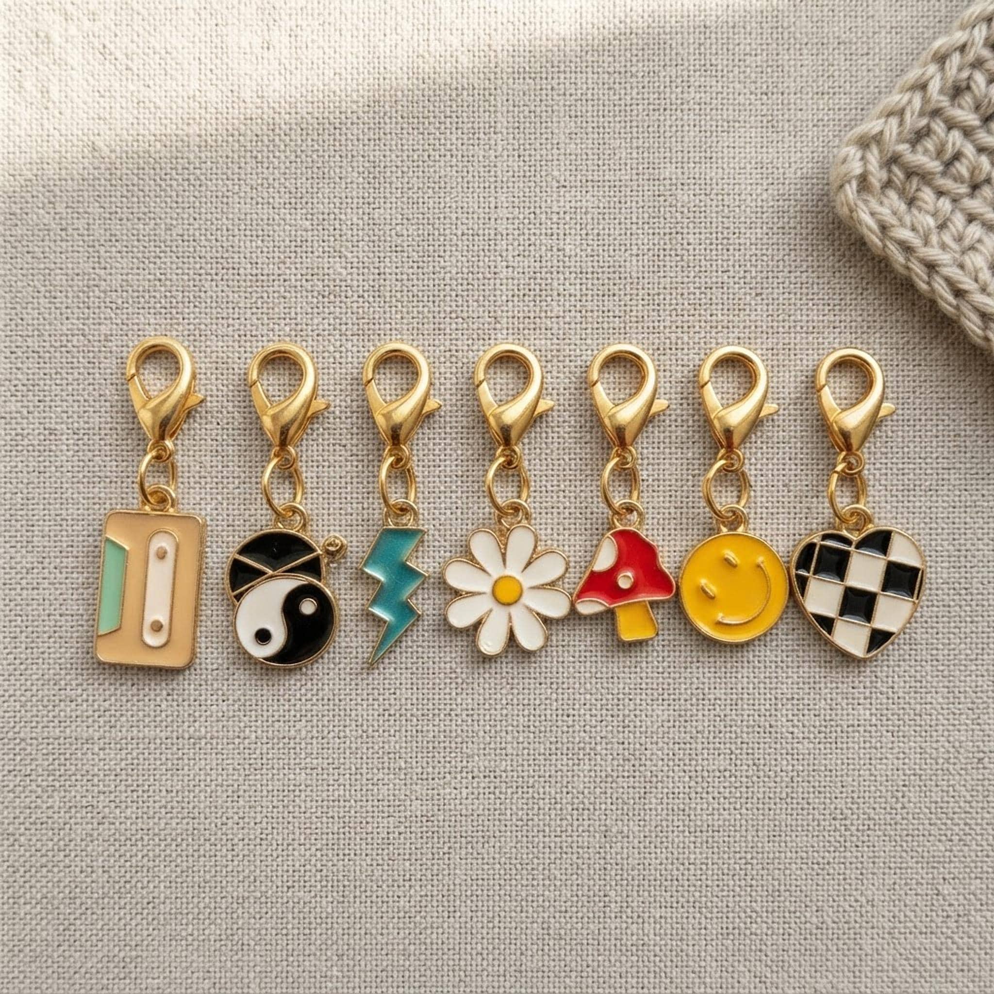 Close up of 8 enamel stitch markers with gold clasps on a linen surface.