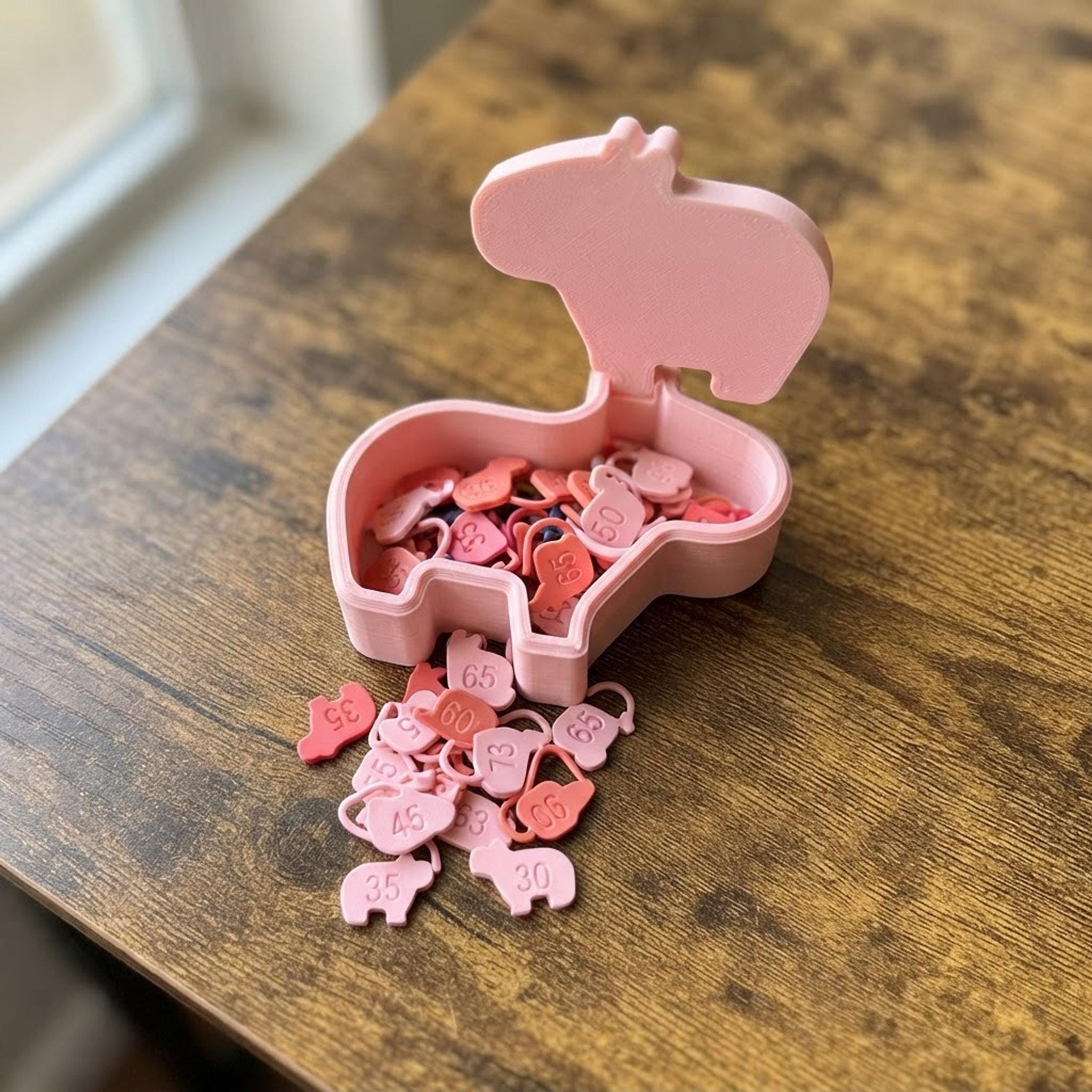A cute pink capybara-shaped container with stitch markers spilling out on a wooden surface.