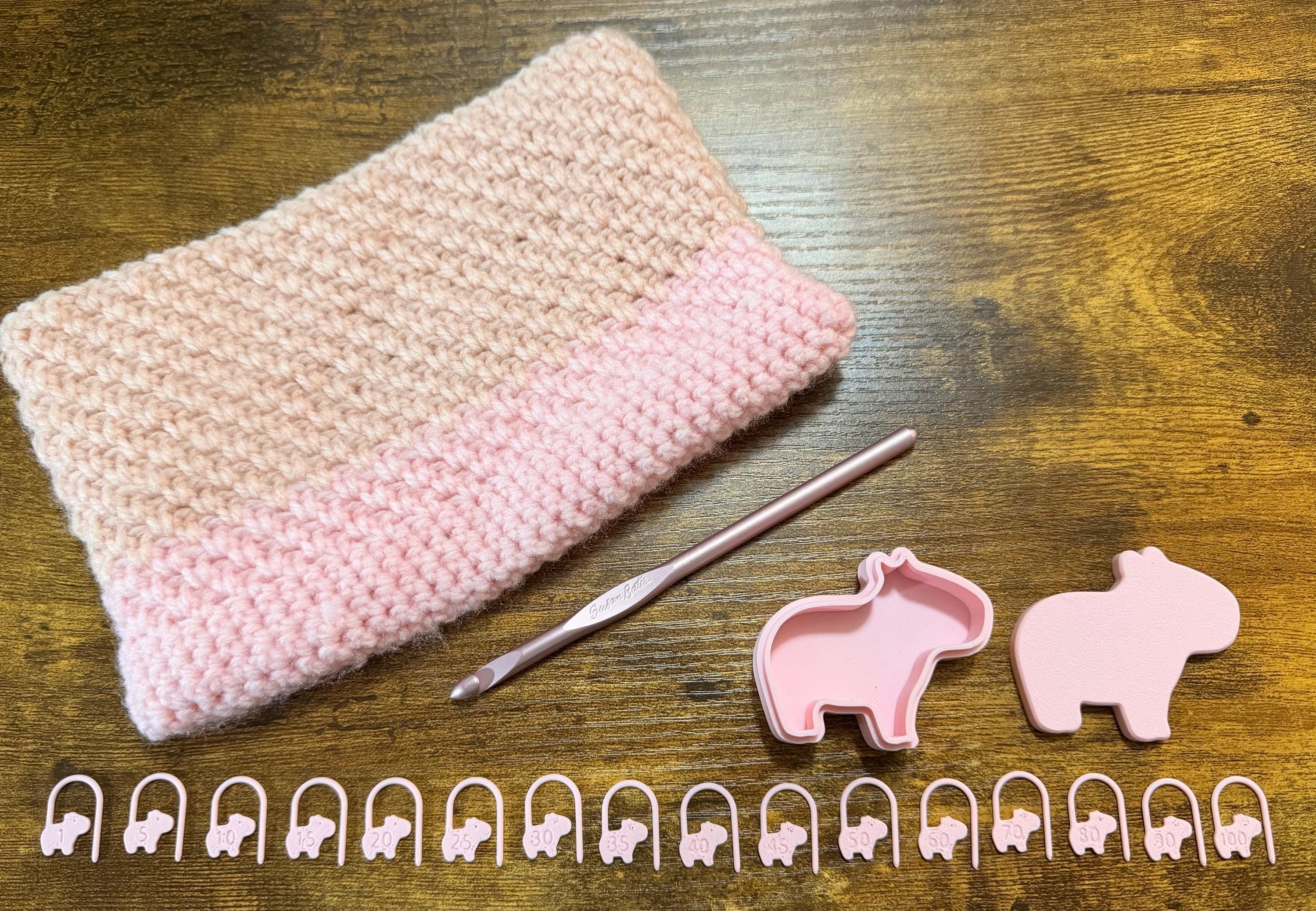 A crochet hook, a folded pink and tan crocheted piece, two pink capybara shapes, and a row of sixteen numbered pink capybara stitch markers lie on a wooden surface.