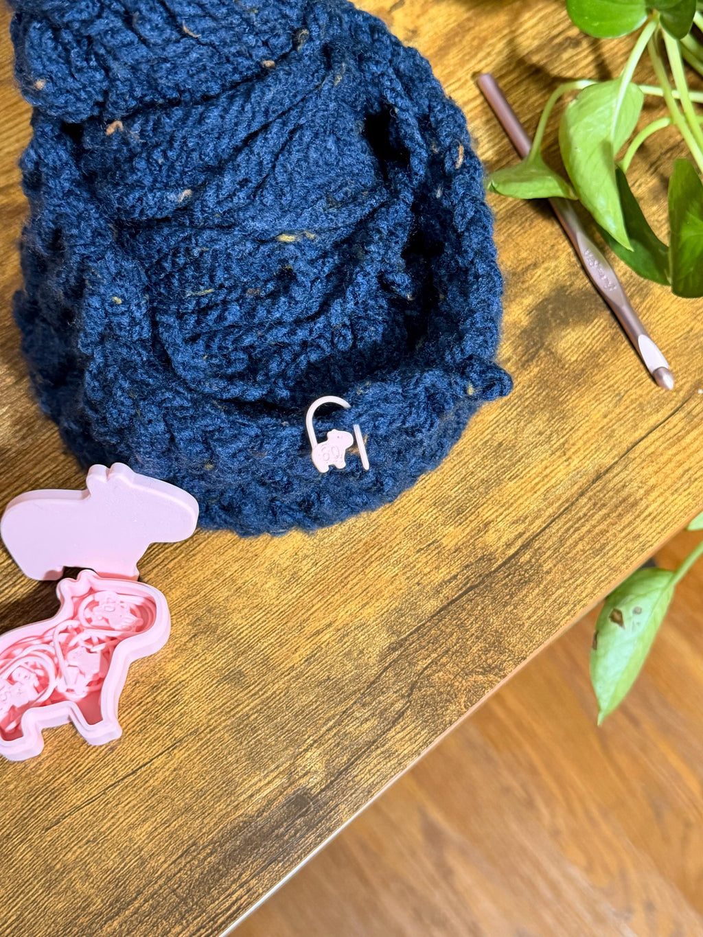 A pink capybara stitch marker is attached to a textured dark blue crocheted item on a wooden table, next to a pink case, a crochet hook, and a plant.