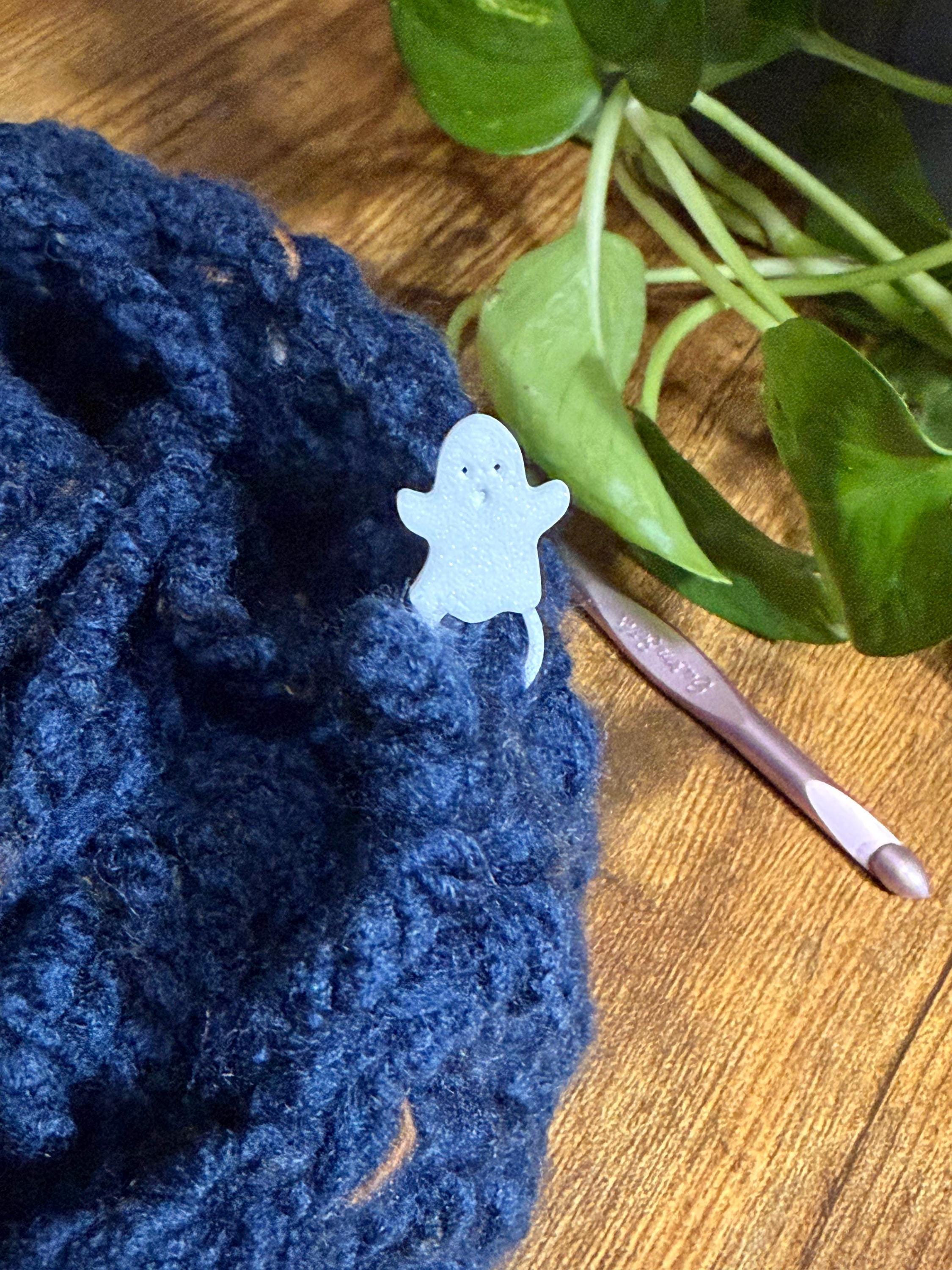 A white ghost stitch marker attached to dark blue crocheted fabric next to a pink crochet hook on a wooden surface.