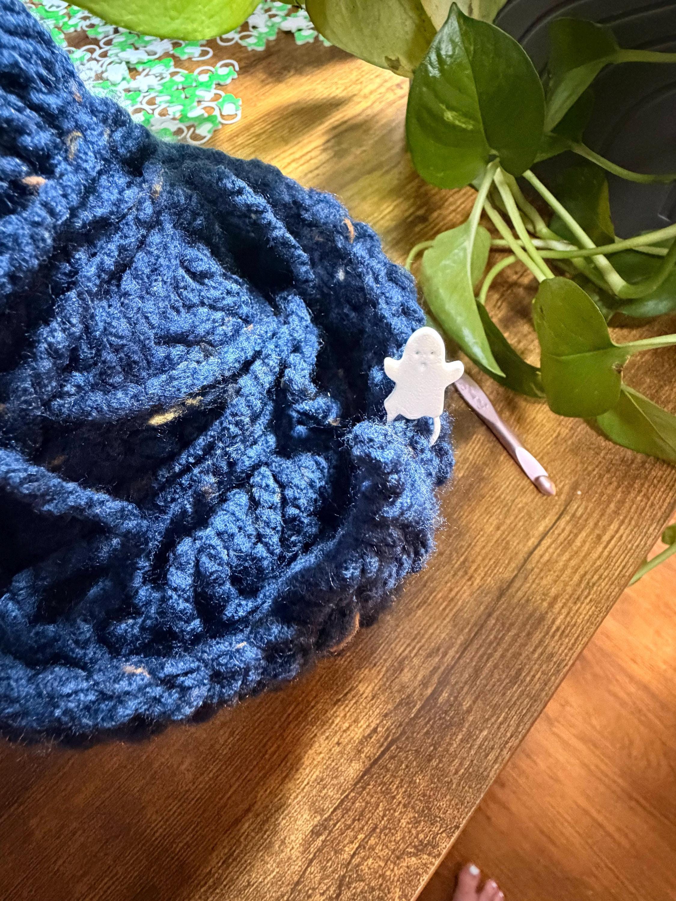 A white ghost stitch marker secured in a ball of textured dark blue yarn on a wooden tabletop with green plant leaves nearby.