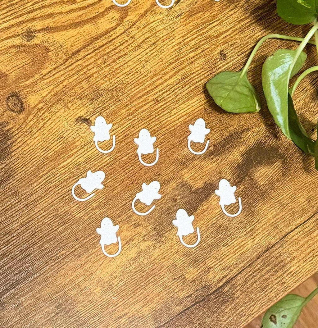 Eight white ghost-shaped stitch markers arranged in a cluster on a wooden surface with green plant leaves in the upper right.