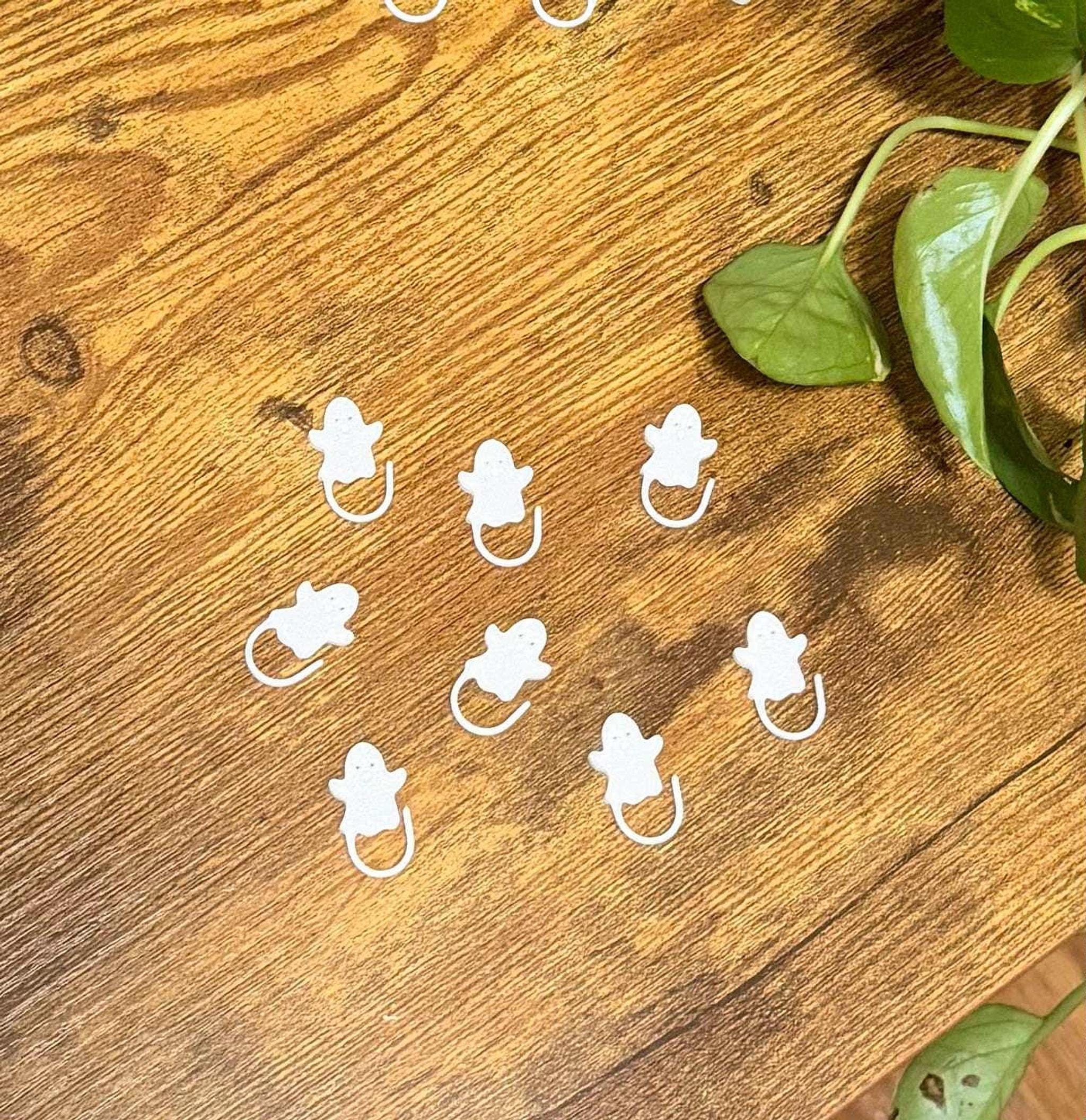 Eight white ghost-shaped stitch markers arranged in a cluster on a wooden surface with green plant leaves in the upper right.