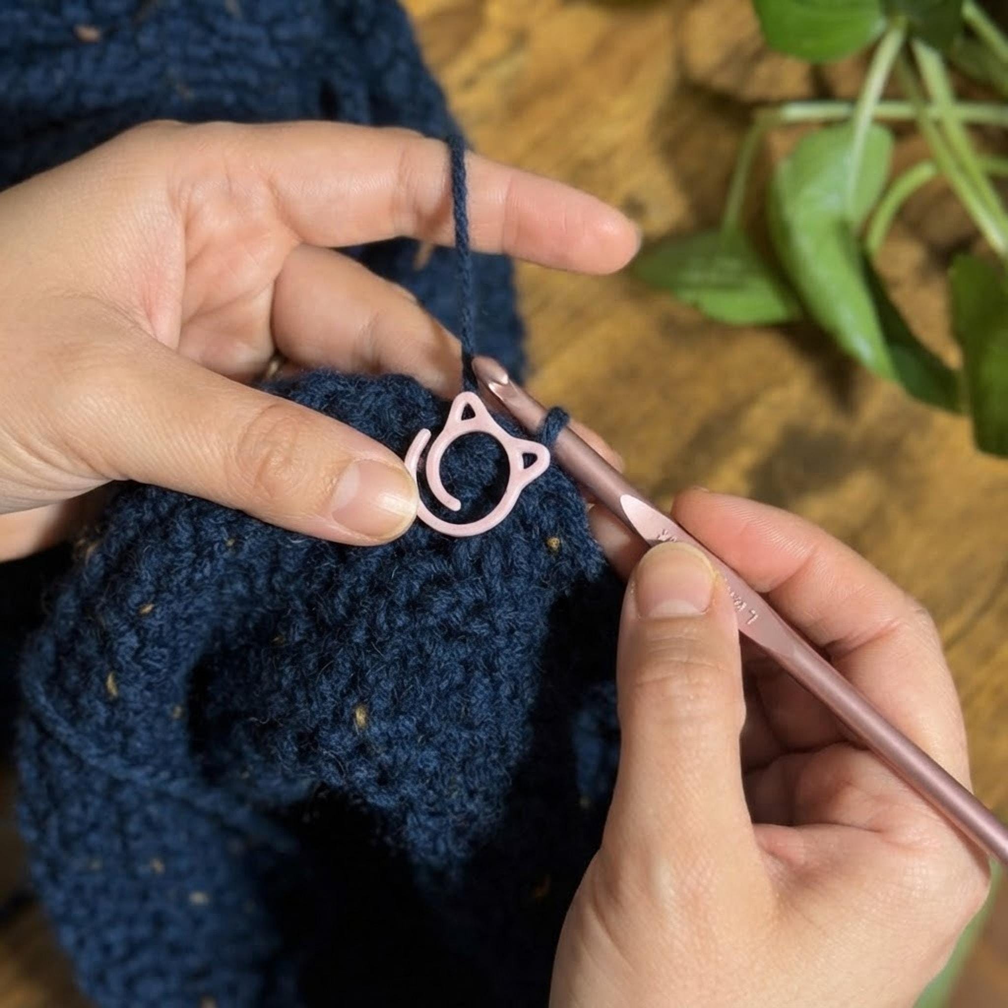 Hands crocheting with a pink cat stitch marker attached to the yarn work for scale.