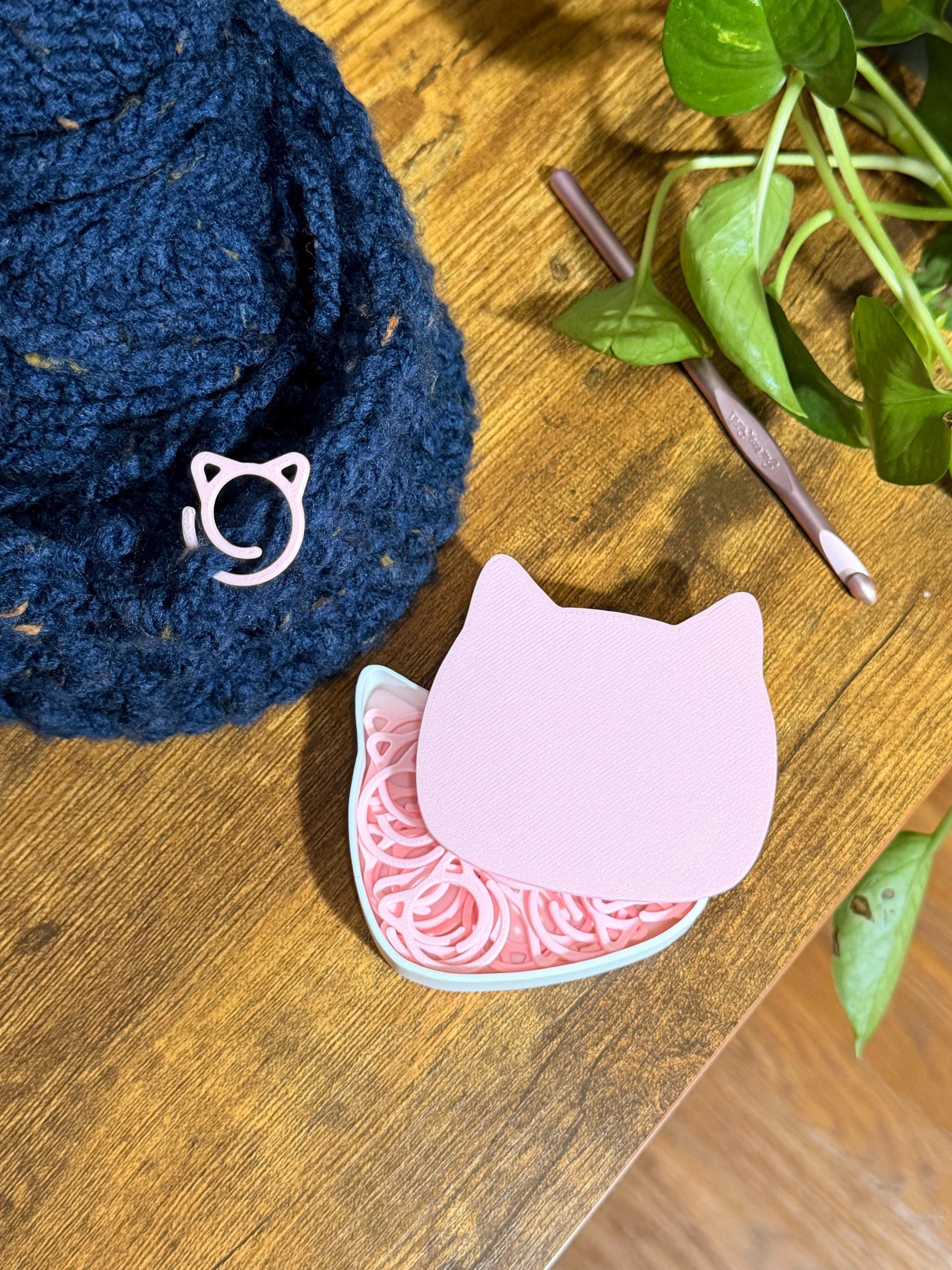 A pink cat-shaped tin holding pink stitch markers sits on a wooden surface next to a crocheted blue hat with a single pink cat stitch marker attached and a crochet hook.