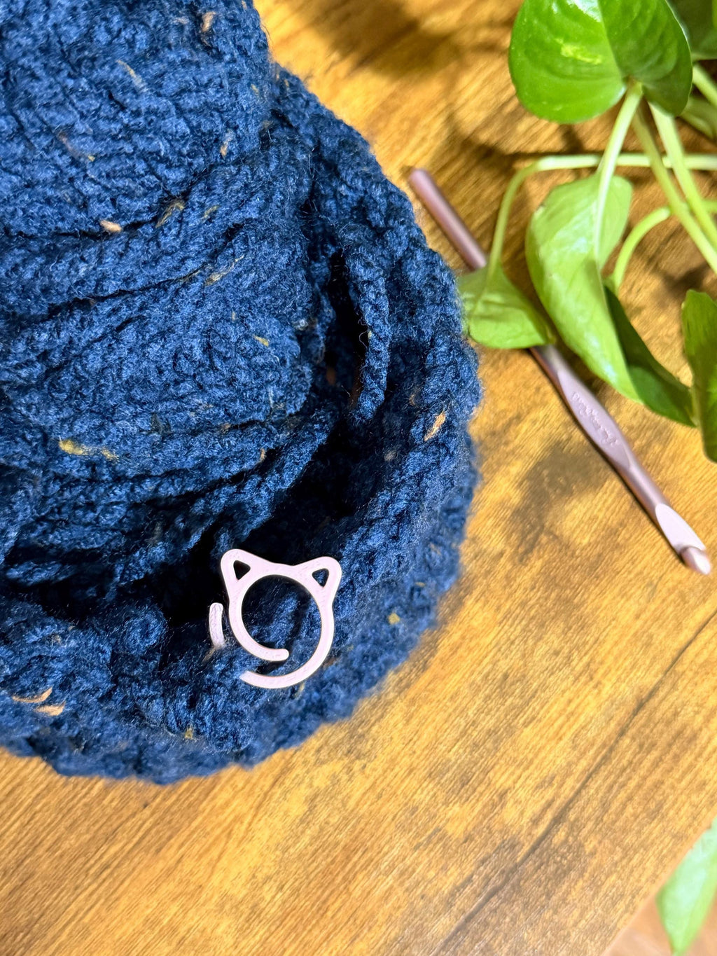 A close-up of a crocheted dark blue hat on a wooden table, featuring a single pink cat-shaped stitch marker attached to the fabric near a crochet hook and a plant.