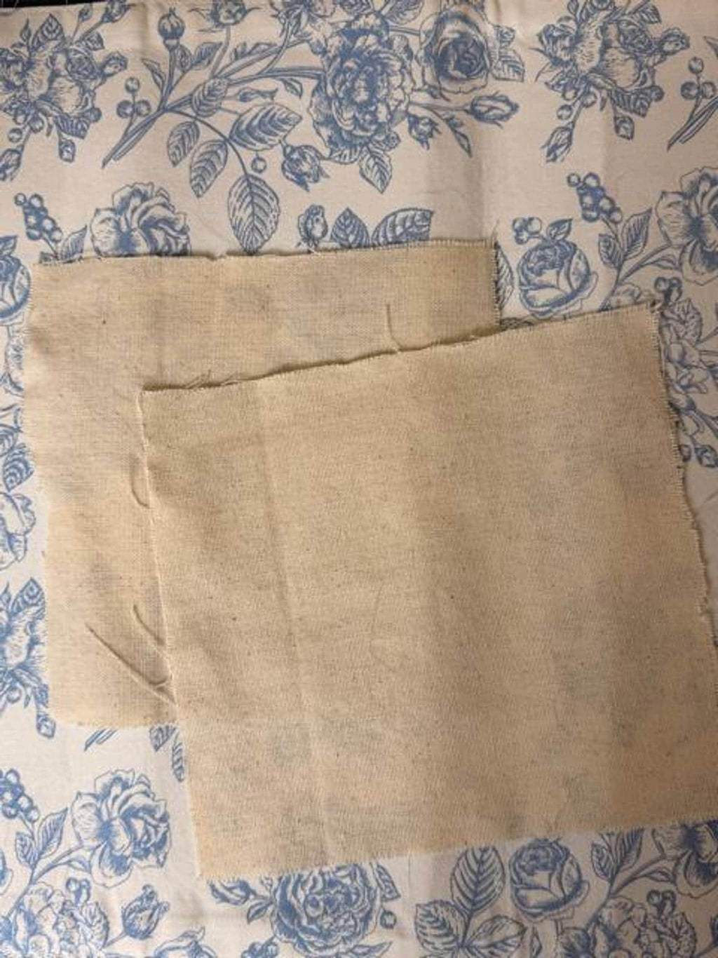 Two squares of beige monks cloth fabric are laid flat on a blue and white floral patterned background.