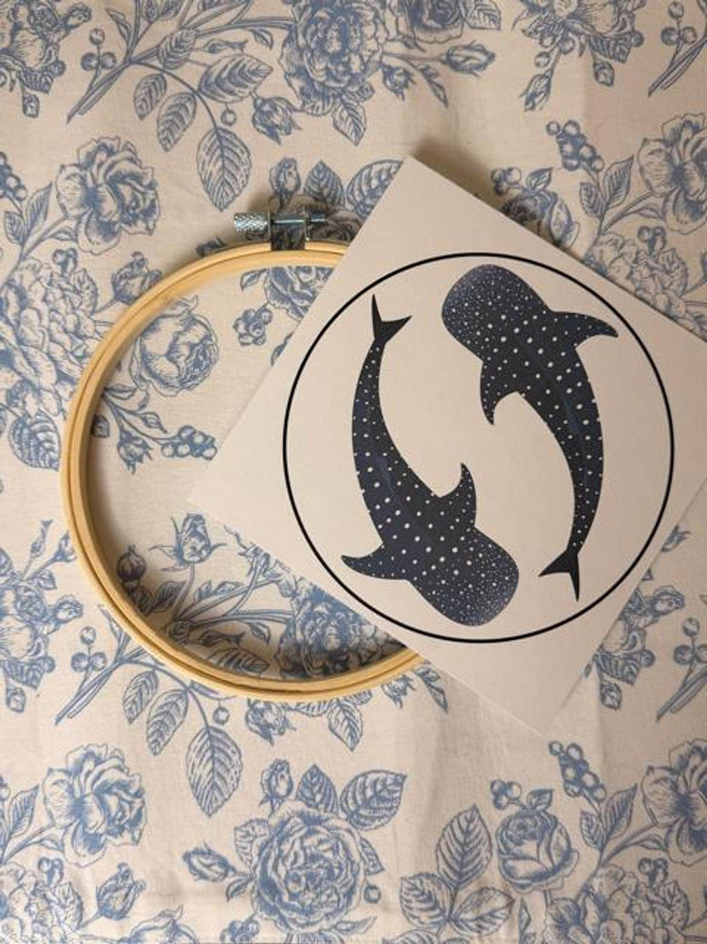 A wooden embroidery hoop and a paper pattern showing two whale sharks inside a circle lie on a floral surface.