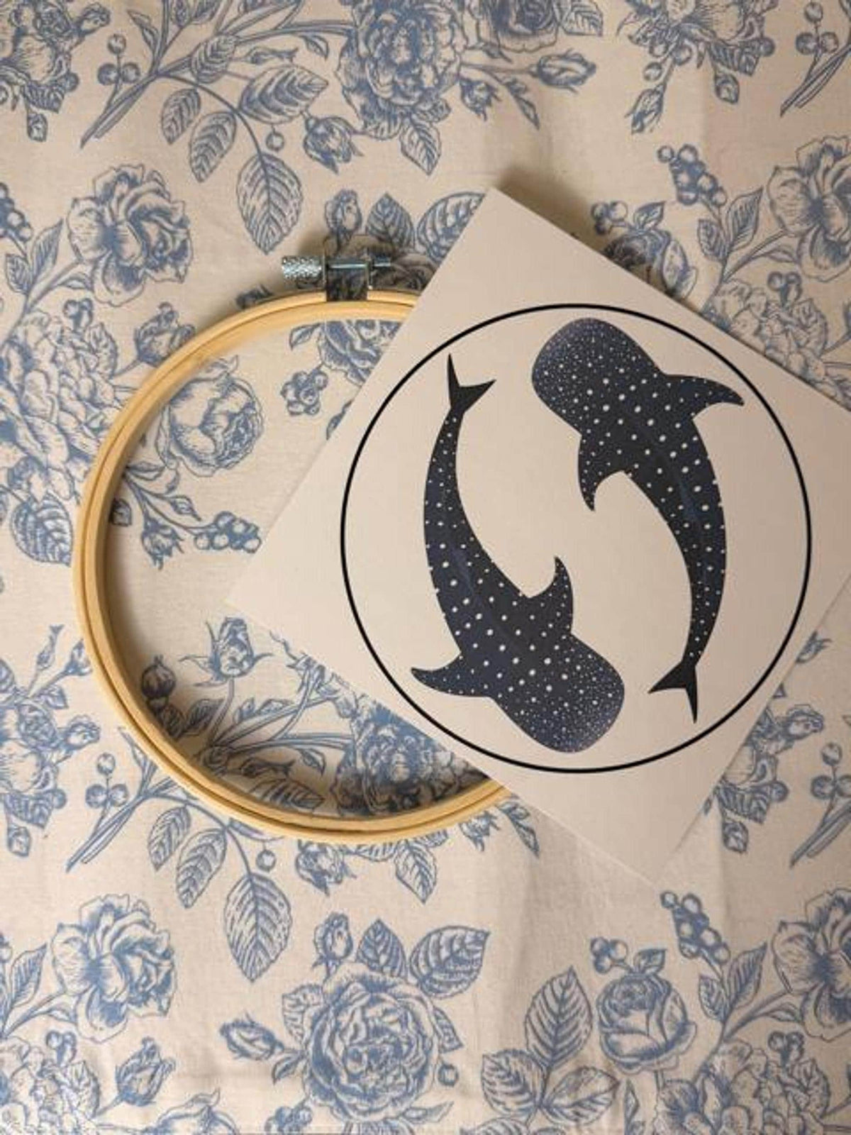 A wooden embroidery hoop and a paper pattern showing two whale sharks inside a circle lie on a floral surface.