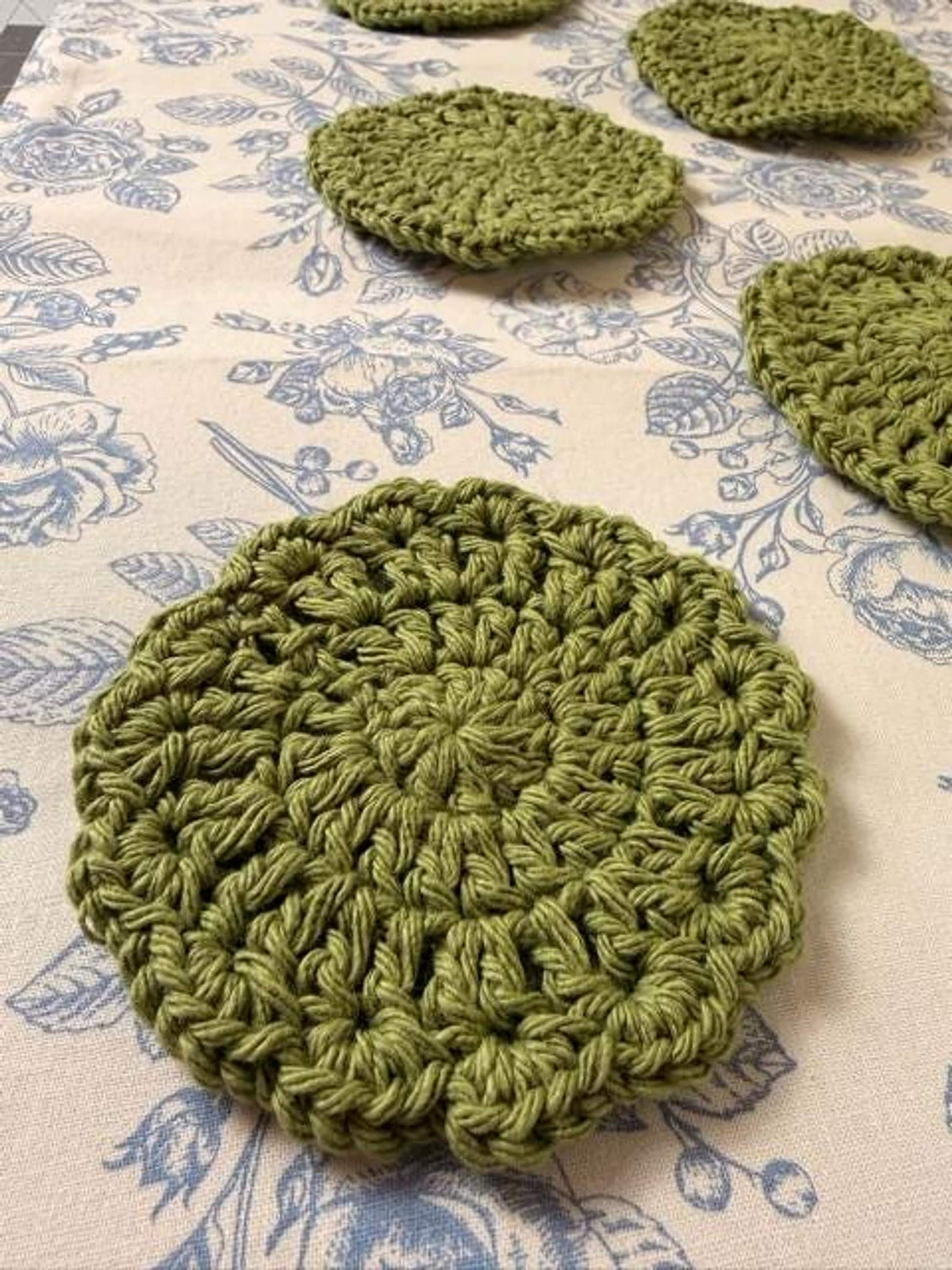 Several round olive green crochet coasters displayed in a staggered pattern on a blue floral fabric.