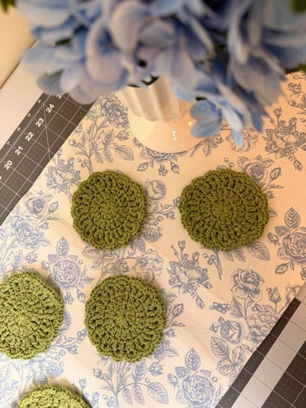 Five round olive green crochet coasters arranged on a blue floral table runner near a vase of blue flowers.