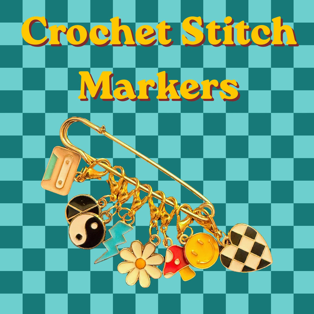 A large gold safety pin holds seven enamel charms including a cassette tape, yin-yang, lightning bolt, daisy, mushroom, smiley face, and checkered heart against a teal checkered background with yellow text reading Crochet Stitch Markers.