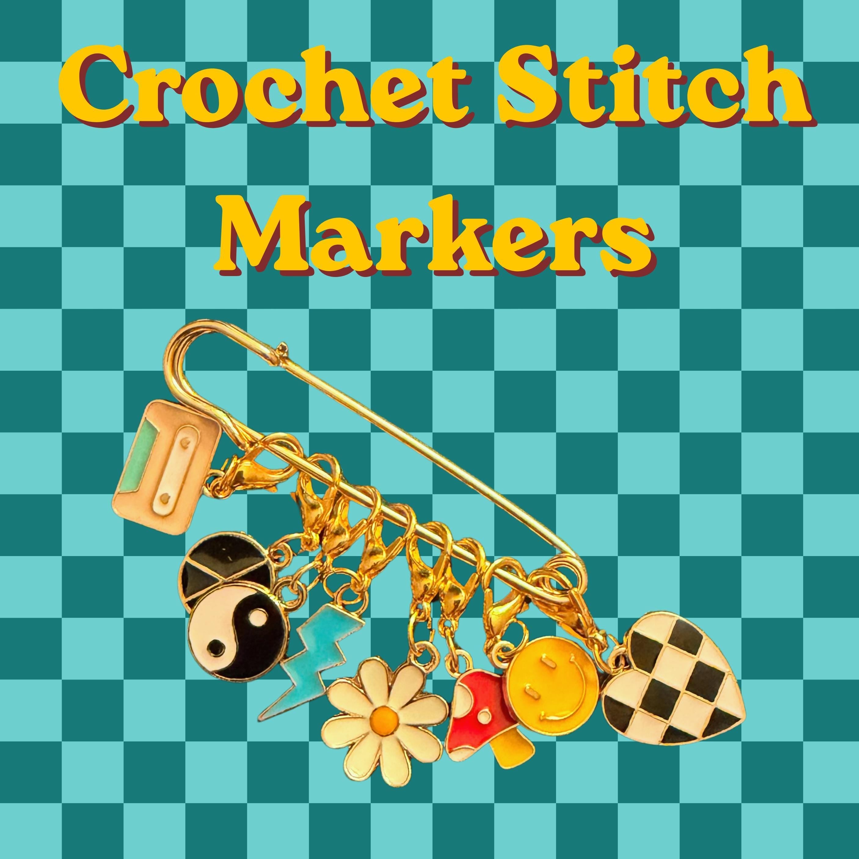 A large gold safety pin holds seven enamel charms including a cassette tape, yin-yang, lightning bolt, daisy, mushroom, smiley face, and checkered heart against a teal checkered background with yellow text reading Crochet Stitch Markers.