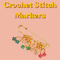 A large gold safety pin holding multiple smaller gold-toned stitch markers with colorful rhinestone flower charms against a solid peach background, with yellow text above reading Crochet Stitch Markers.