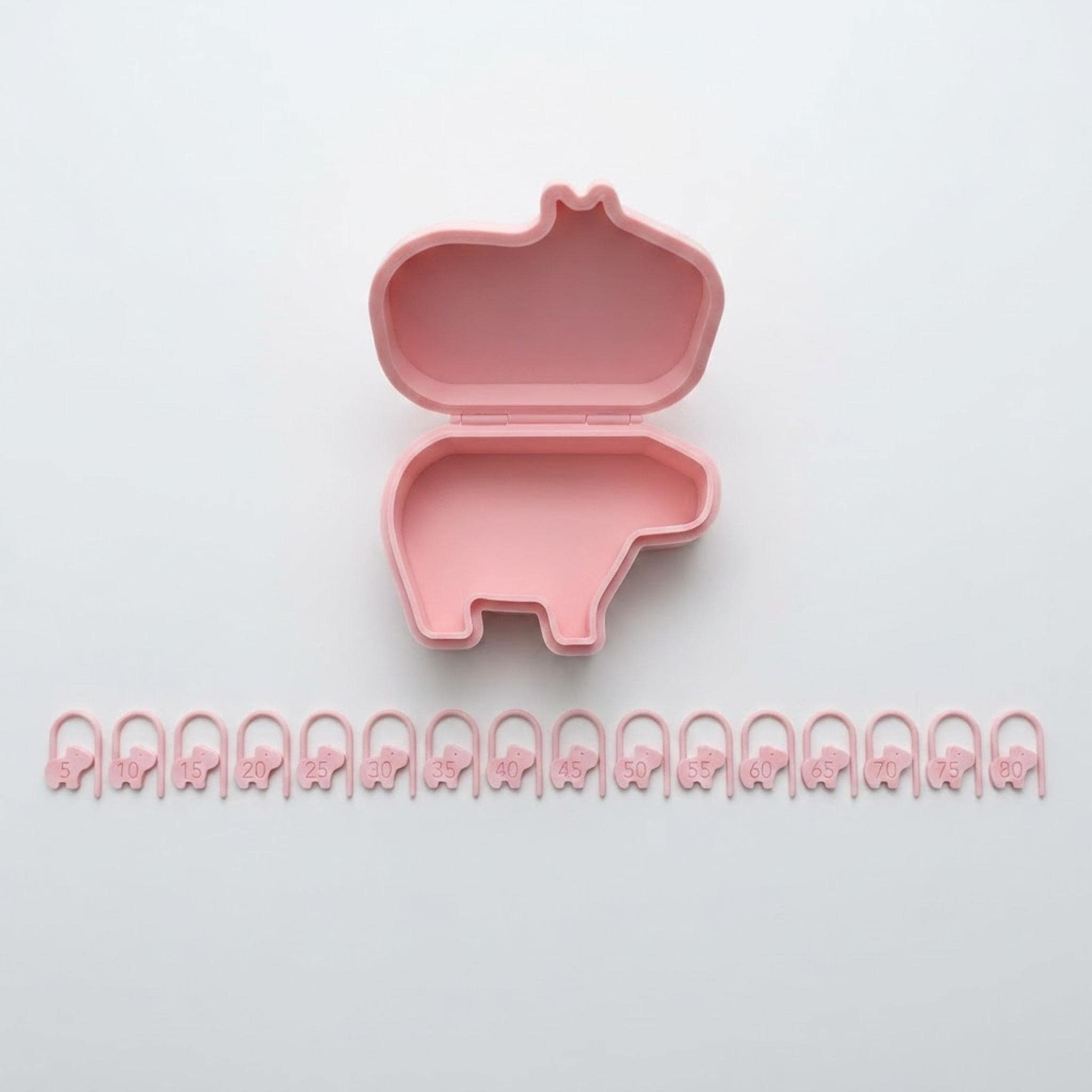 A complete set of 16 pink capybara stitch markers with numbers laid out flat next to their capybara-shaped storage case.