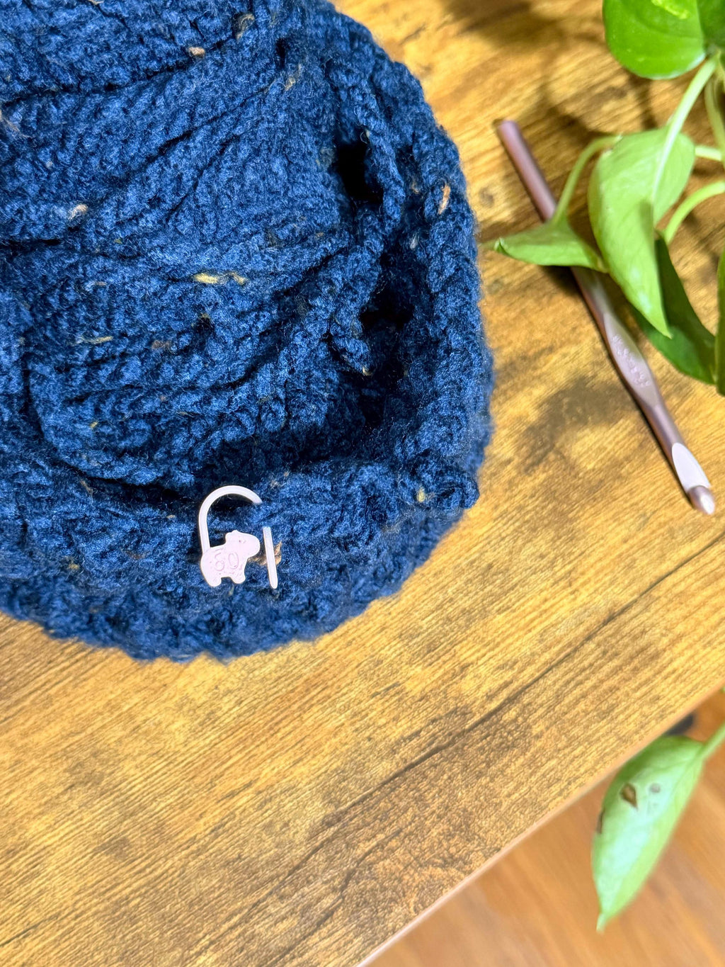 Close-up of a pink capybara stitch marker numbered 50 attached to a dark blue crocheted fabric resting on a wooden table near a crochet hook.