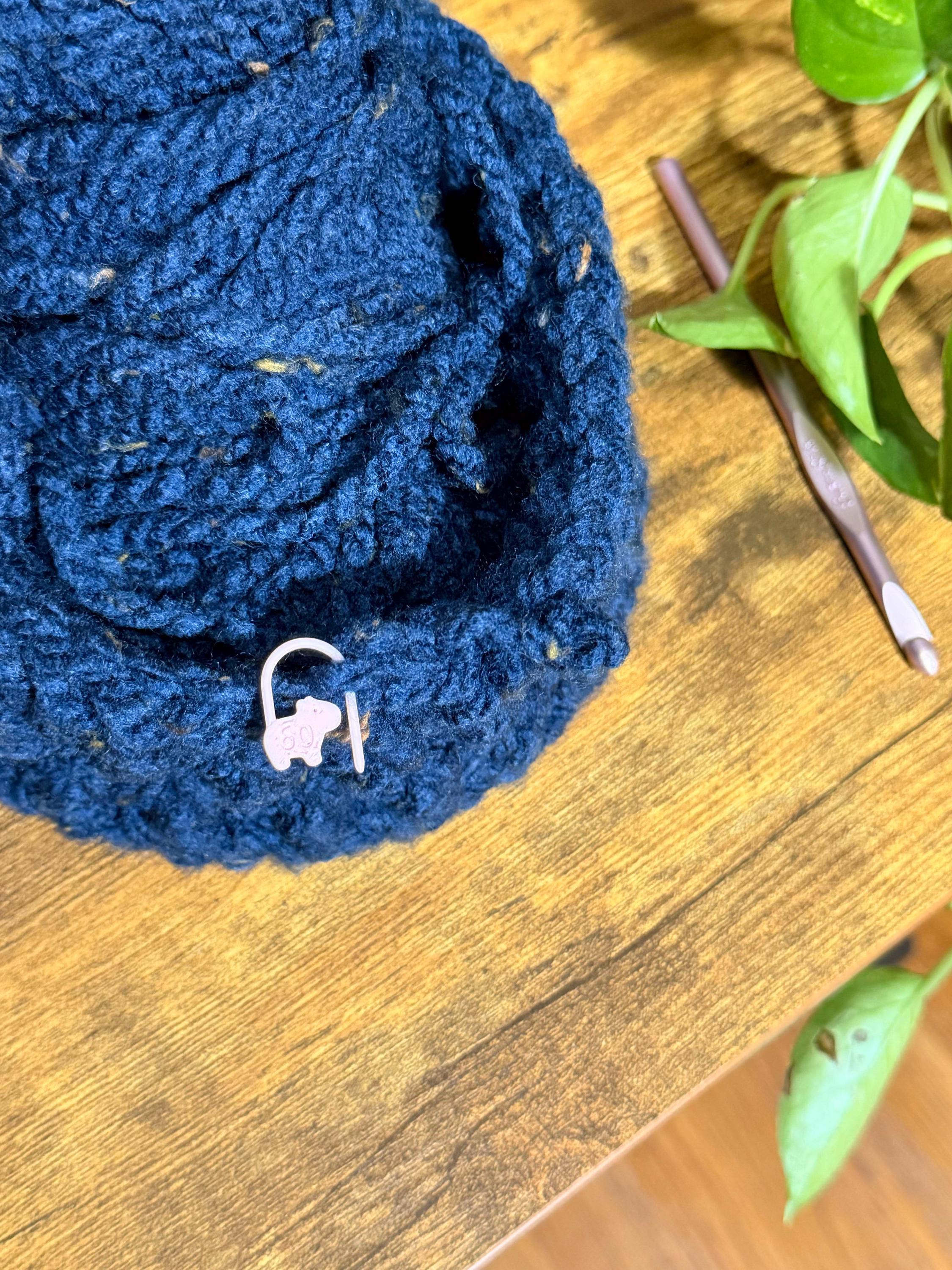 Close-up of a pink capybara stitch marker numbered 50 attached to a dark blue crocheted fabric resting on a wooden table near a crochet hook.