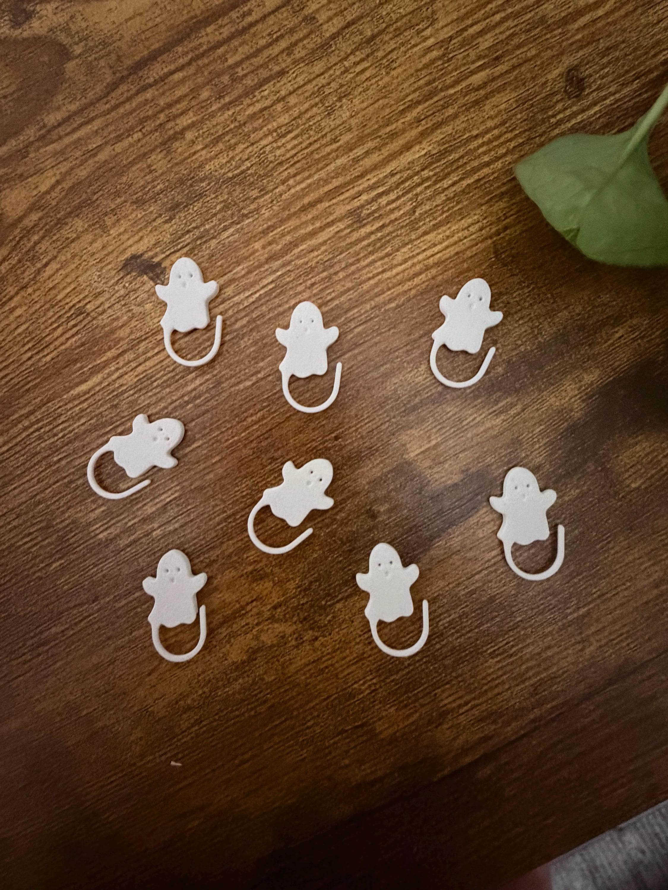 Eight white ghost stitch markers scattered on a wooden table with a single green leaf near the top right.
