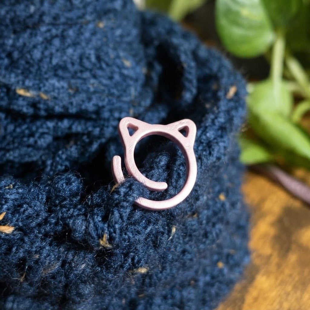 Macro view of a pink cat stitch marker attached to textured navy blue crochet yarn.