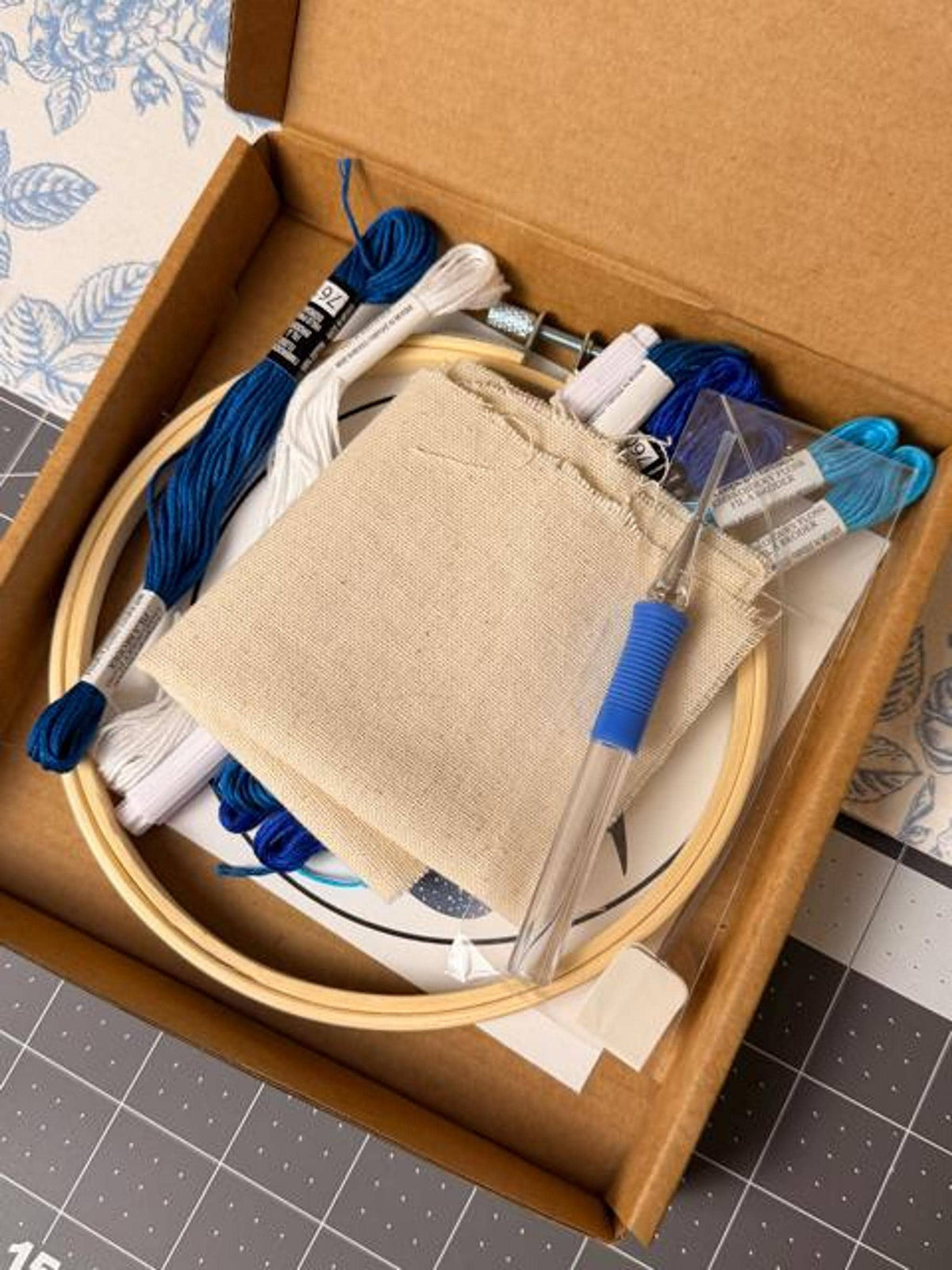 Inside the craft box are a wooden embroidery hoop, blue and white embroidery floss, monks cloth, and a punch needle tool.