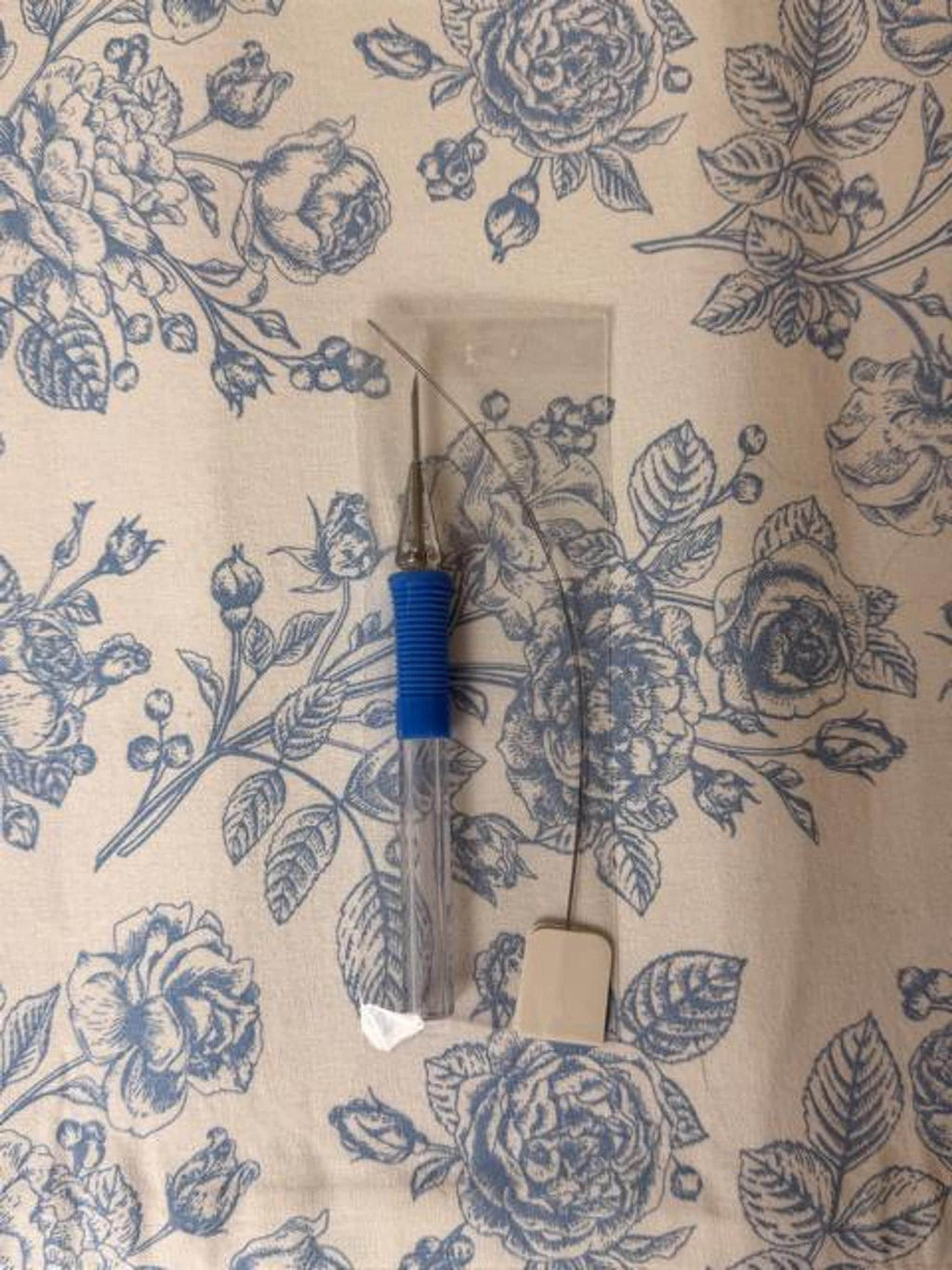 A punch needle tool with a blue textured grip and a wire threader lie on a floral fabric.