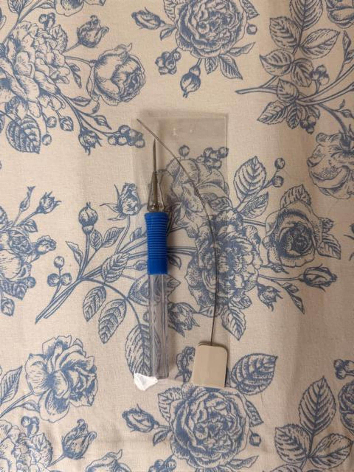 A punch needle tool with a blue textured grip and a wire threader lie on a floral fabric.