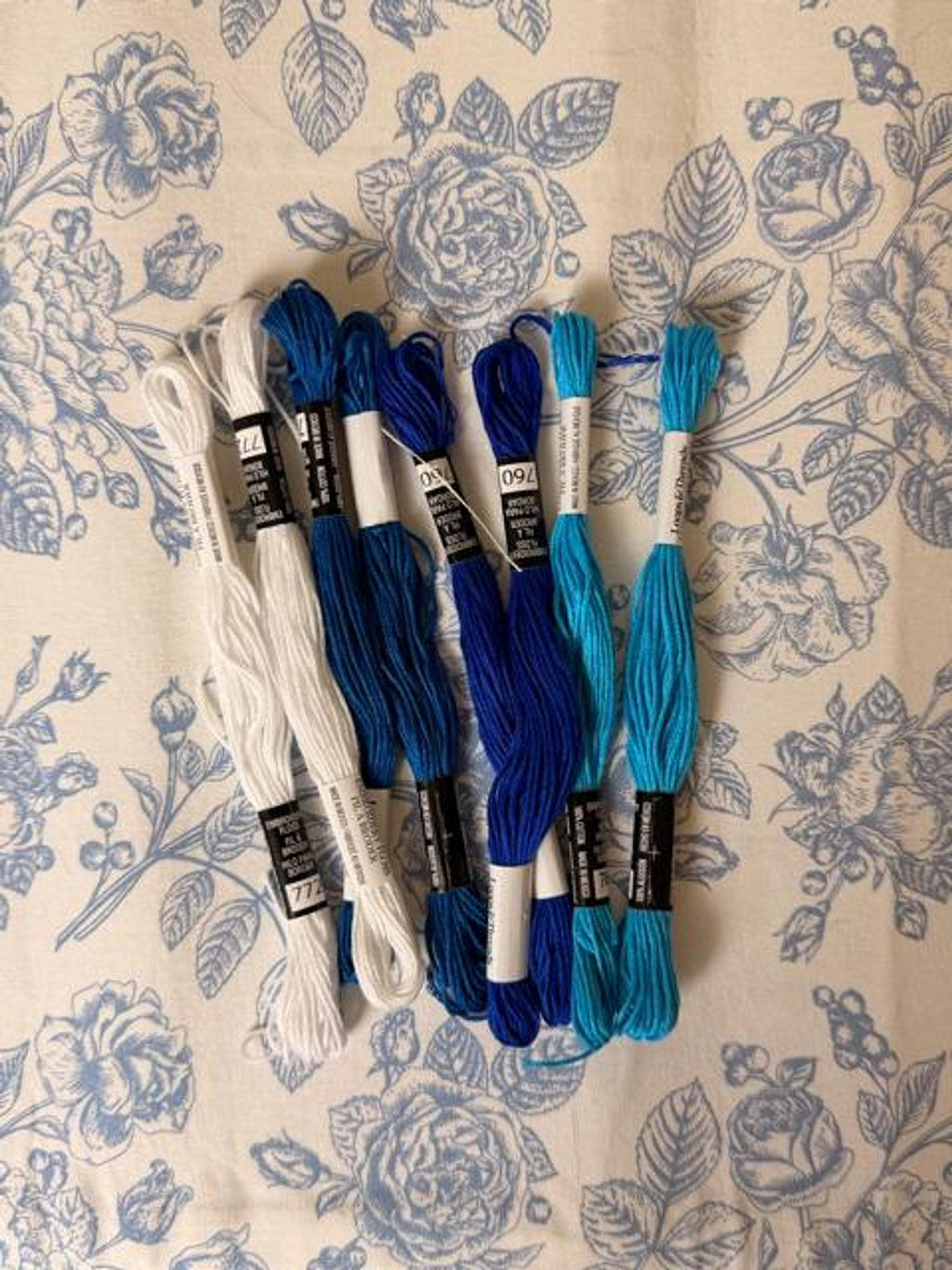 Skeins of embroidery floss in shades of white, dark blue, royal blue, and turquoise are arranged on a floral fabric.