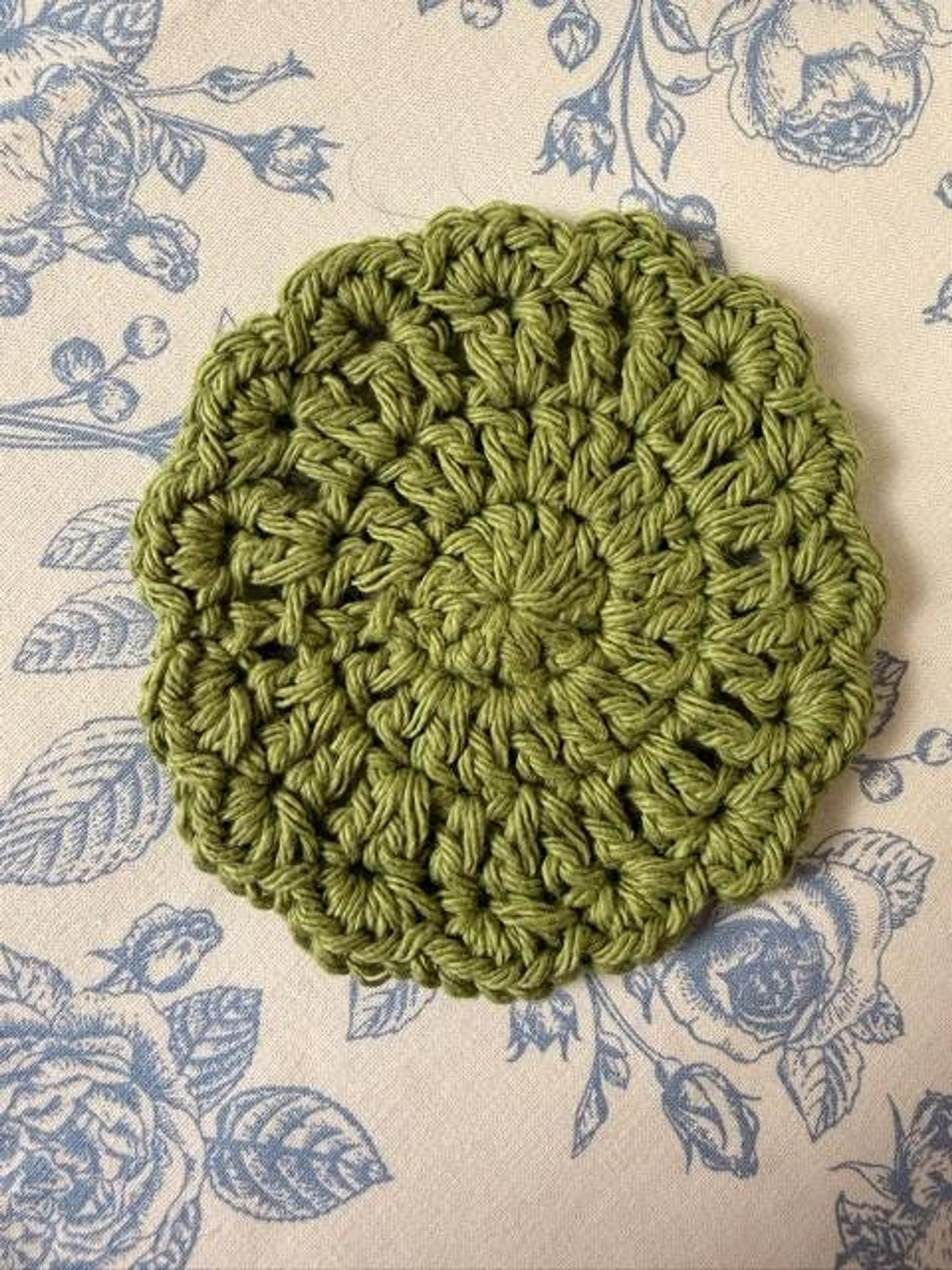 A single round olive green crochet coaster laid flat on a blue floral tablecloth.