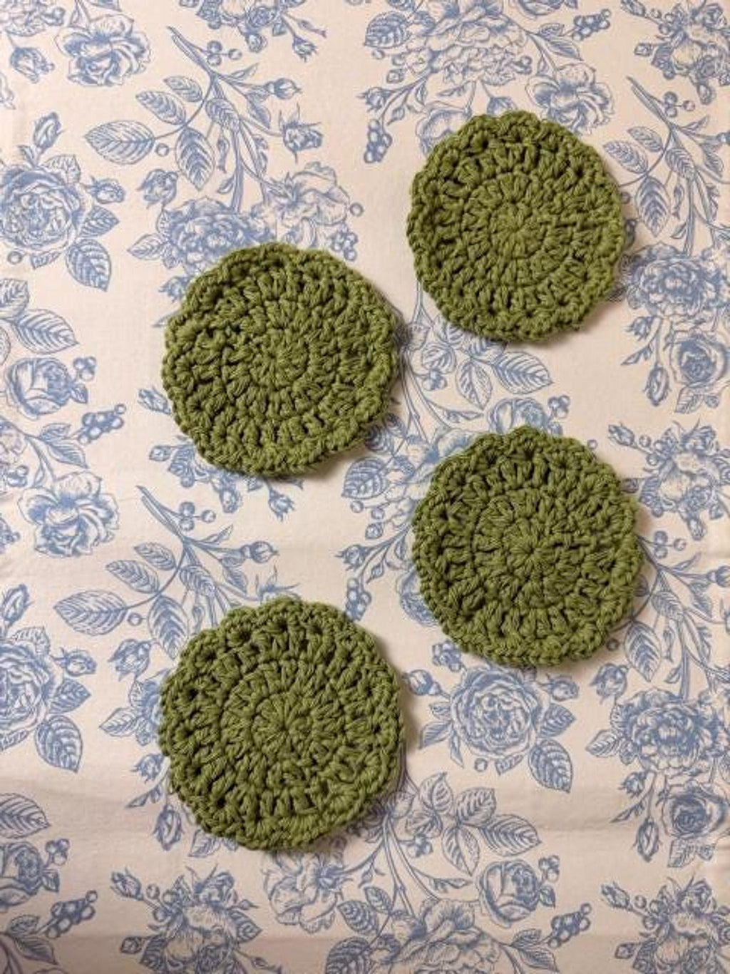 Four round olive green crochet coasters arranged in a square formation on a blue and white floral fabric.