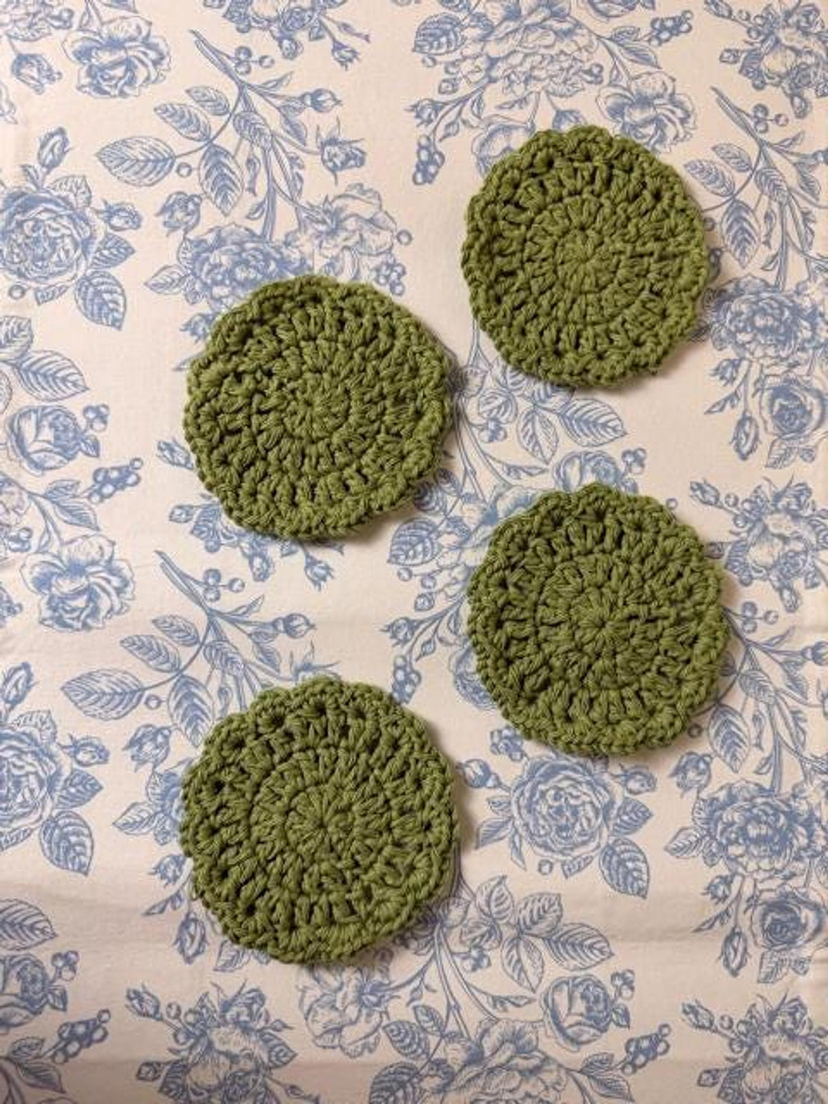 Four round olive green crochet coasters arranged in a square formation on a blue and white floral fabric.
